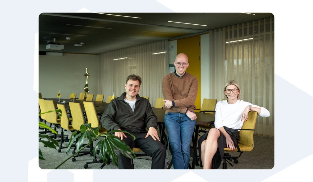 Drie Azumuta teamleden zitten en staan samen in een lichte vergaderruimte met gele stoelen en een modern design. Ze glimlachen naar de camera en brengen een vriendelijke en professionele sfeer over.
