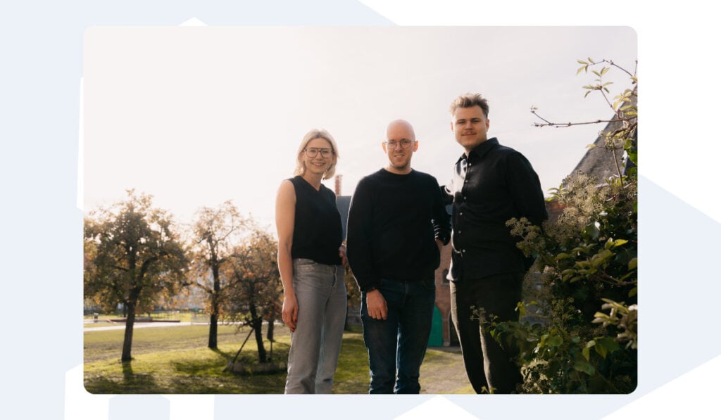 "Three members of the Azumuta team stand together outdoors in soft sunlight. A woman in glasses and a black sleeveless top stands on the left, Batist in a dark sweater and glasses stands in the middle, and a man in a black shirt stands on the right. Behind them is a garden with trees and greenery."