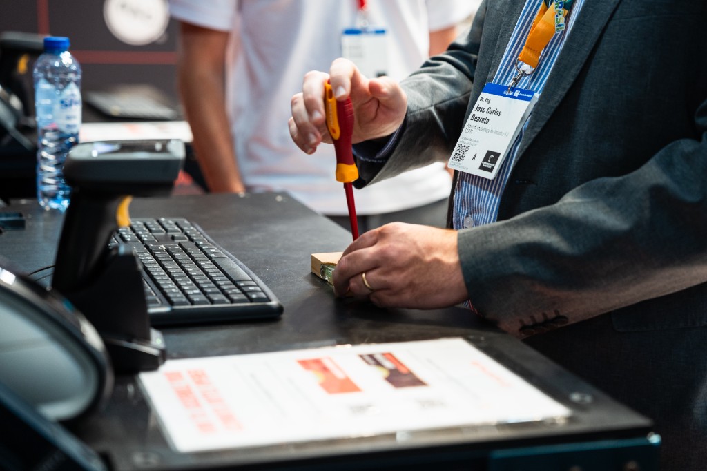 Een beursbezoeker gebruikt een schroevendraaier om een bierflesopener te monteren, stap voor stap begeleid door digitale werkinstructies op de Azumuta-stand.