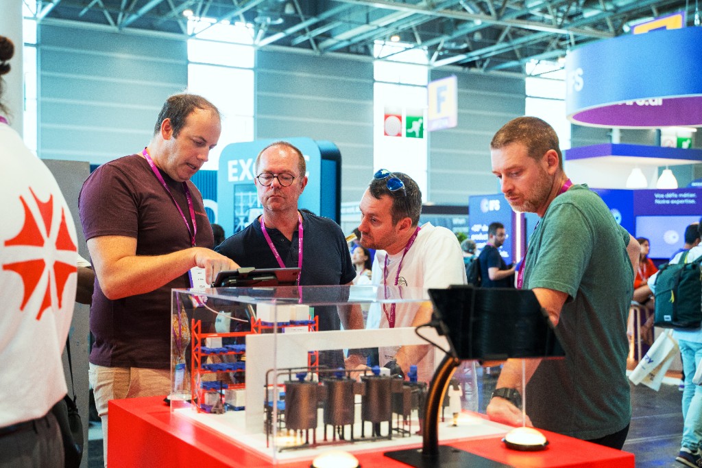 Bezoekers verzameld rond een demo van een miniatuurproductielijn op de Azumuta-stand tijdens Tech For Industry Show in Parijs.