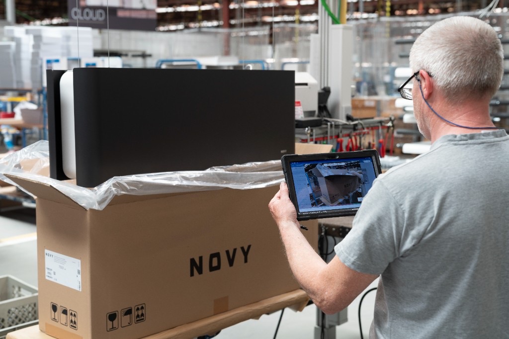 An operator at the Novy line follows Azumuta digital work instructions on a rugged handheld tablet while checking a packed product against the on-screen steps.